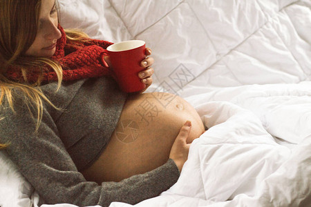 在床上的年轻孕妇拿着茶杯握图片