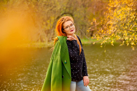 穿着可爱的蓝色毛衣心胸细小的红发美女在秋天公园冷瀑湖附近拿着绿大衣图片