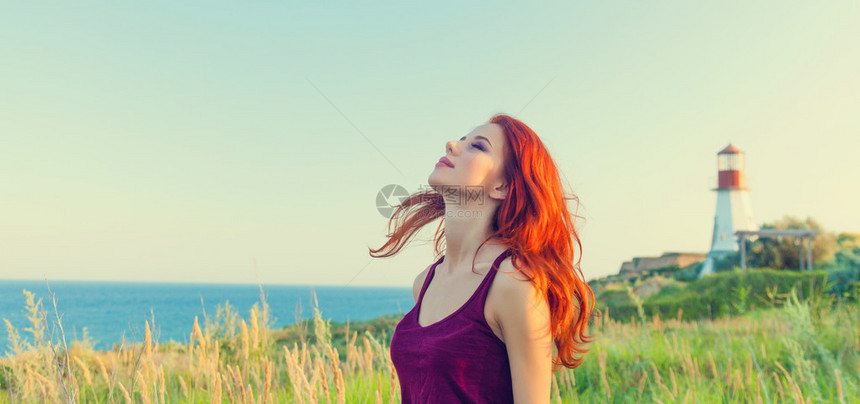 背景中灯塔和蓝色大海附近的年轻红发女孩图片