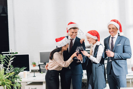在圣塔帽子中快乐的年轻同事喝酒和在工作场所庆祝圣诞日图片