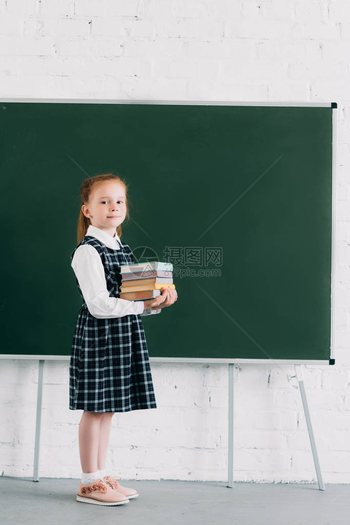 可爱的小女学生拿着一堆书看摄影机站在黑板旁图片