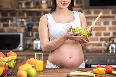 微笑的年轻孕妇在厨房里拿着装有健康蔬菜沙拉的图片