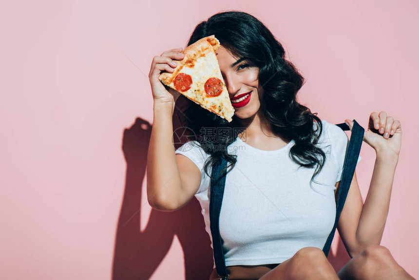 在粉红色背景上用一块披萨遮着眼睛的美丽的微笑女图片