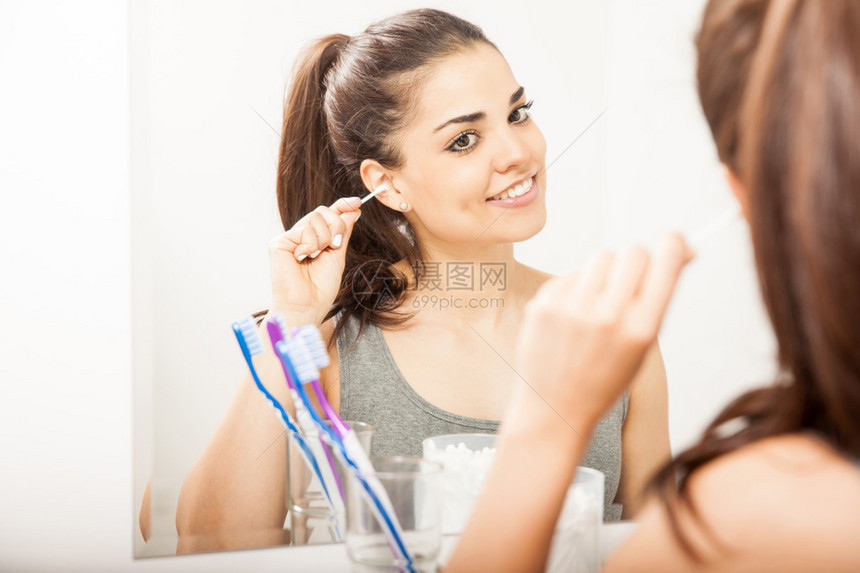 美丽的年轻女子站在浴室的镜子前用棉签清洁耳朵图片