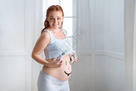 幸福的孕妇肚子上戴耳机家里戴着微笑符号的图片