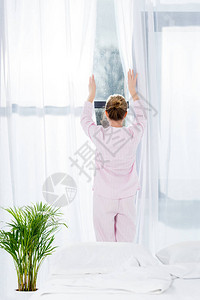 穿着睡衣的女人早上拉开窗帘的后视图背景图片