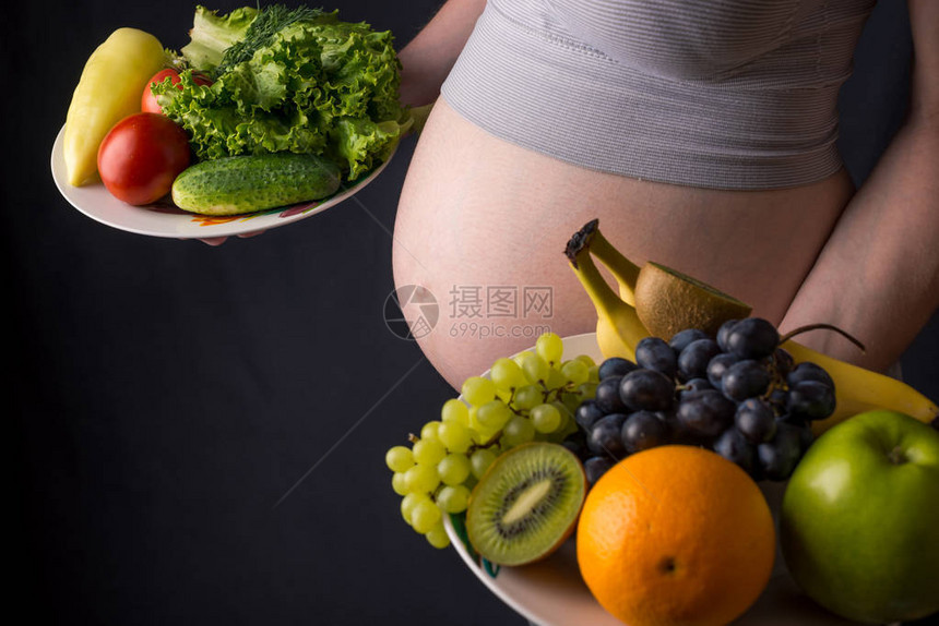 一个有肚子的孕妇手里拿着一盘水果和蔬菜孕期体重控制和健