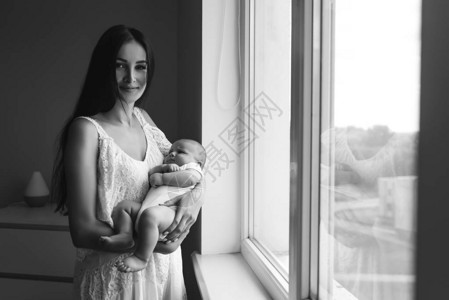 美丽的年轻母亲在家中的窗户旁边携带婴儿并观看相机的黑白镜头图片