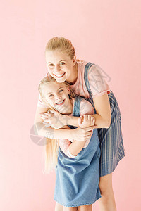 穿着蓝裙子的年轻快乐母亲和女儿拥抱并看着在粉红色上图片