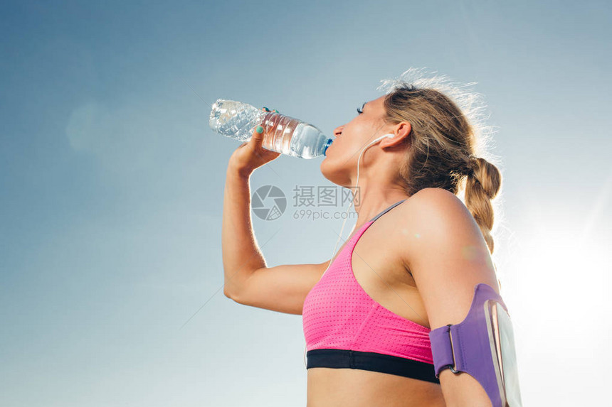 青年运动女运动员用耳机和智能手机在臂带运动个案中从瓶装饮用水到图片