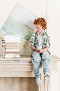 沉思的小男孩坐在一堆书旁的壁炉上图片