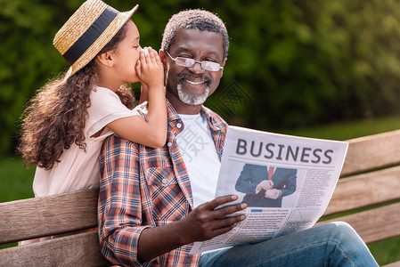 非洲裔美国女孩在她的祖父坐在长凳上阅读商业报图片