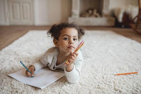 可爱的非洲小女孩用铅笔画躺在家里地毯背景图片