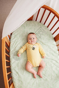 身穿黄色连体衣独自躺在婴儿床靠近窗边的可爱风趣可爱白人小婴儿的画像图片