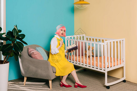 时尚的孕妇被粉色头发缠着笔记本和在婴儿床附近看相机的女人绑起来图片