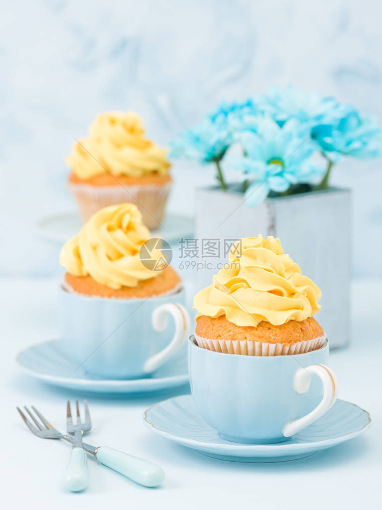 带有黄色奶油装饰的纸杯蛋糕和蓝色柔和背景上复古破旧别致花瓶中的蓝色束图片