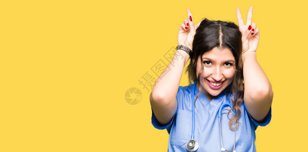 身穿医疗制服的年轻成年女医生用手指头上的兔子耳朵摆出滑稽而疯狂的姿势背景图片