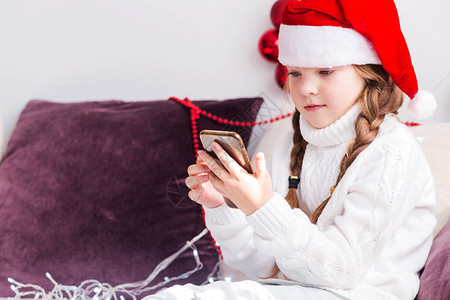 一个穿白色毛衣的小女孩和圣诞老人帽子看手机在她的手中横向照图片