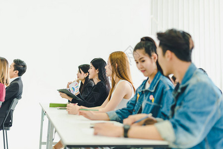 亚洲人参加研讨会并在培训室聆听演讲者的演讲有些人会记笔有人举手询问解说员并在演讲背景