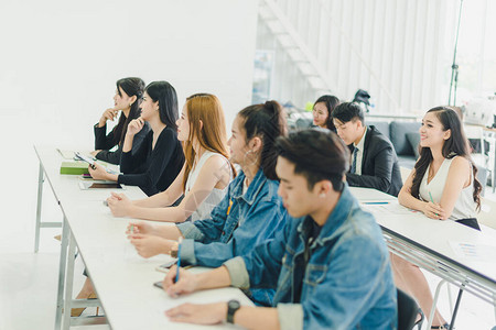 亚洲人参加研讨会并在培训室聆听演讲者的演讲有些人会记笔有人举手询问解说员并在演讲背景