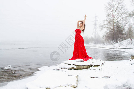 年轻穿长红色裙子的女孩站在雪河背景图片