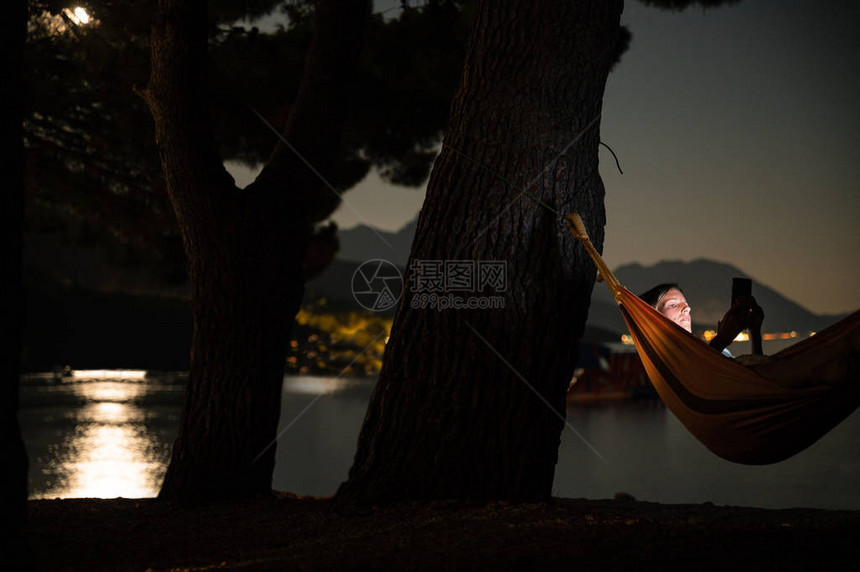 躺在吊床上的年轻女子在晚上的海边浏览她的手机时图片