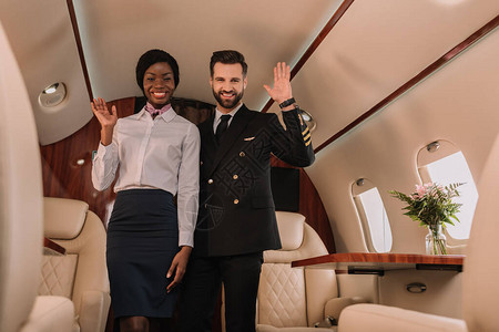 快乐的飞行员和笑的Affican美国空姐挥手握在私人喷气式飞机图片
