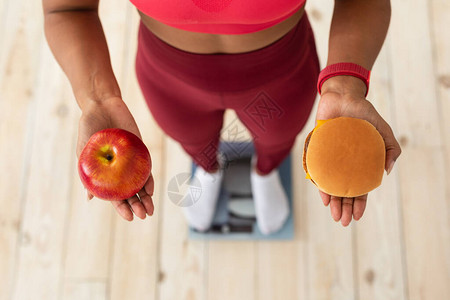 健康饮食和体重损失在汉堡与苹果之间选择黑女孩在家里的重量比重上站立图片