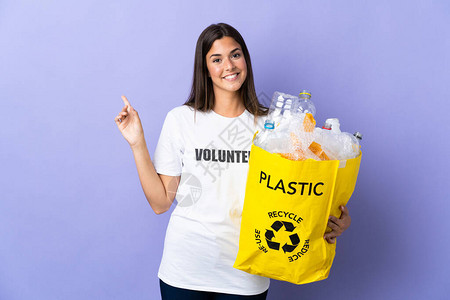 拿着一袋装满塑料瓶的袋子来回收利用的年轻巴西女人在紫色背景中图片