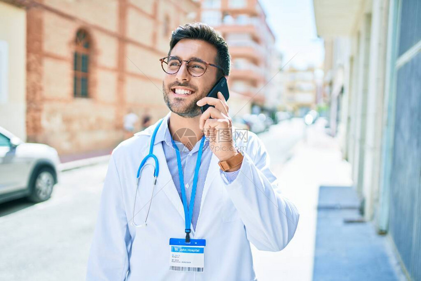 年轻英俊的西班牙裔医生戴着听诊器微笑着开心地站在镇街上用智能手机说话时图片