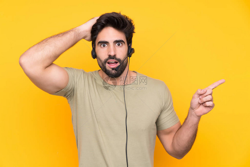 电讯推销员用耳机为孤立的黄色背景工作的人感到惊讶图片