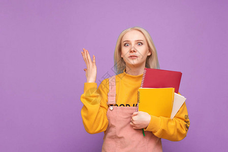 手里拿着书和笔记本的被吸引的情绪化女孩站在紫色的背景上背景图片