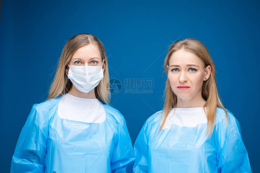 防护概念两位身穿蓝色防护袍的医生左边的女人戴着无菌口罩右边的女人带着害怕的表情看着相机在蓝色上隔图片