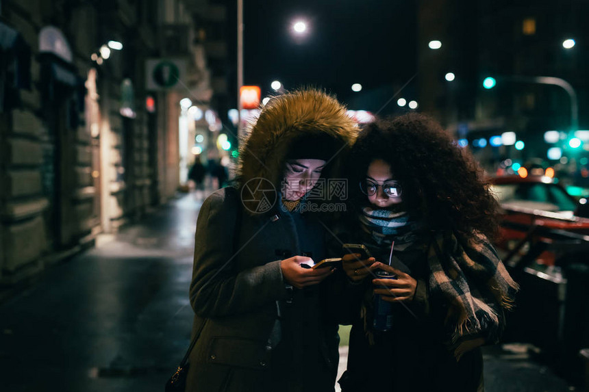 两名多族裔青年妇女夜间在户外使用智能电话Phubbing社交网图片
