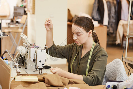 在缝纫机前坐在桌前用针线和针线缝衣的年轻图片