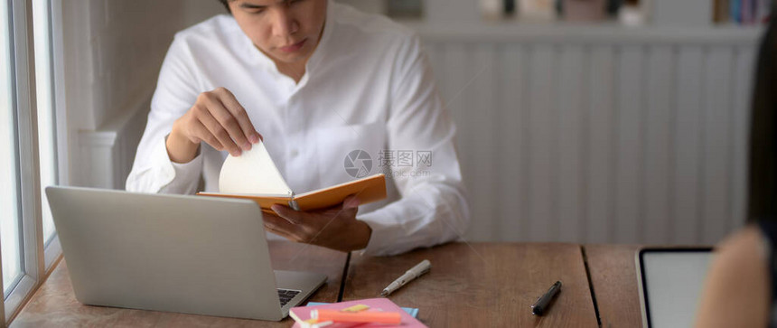 用笔记本电脑和笔记本在图书馆木制书桌上准备考试的年轻男大学生被剪图片