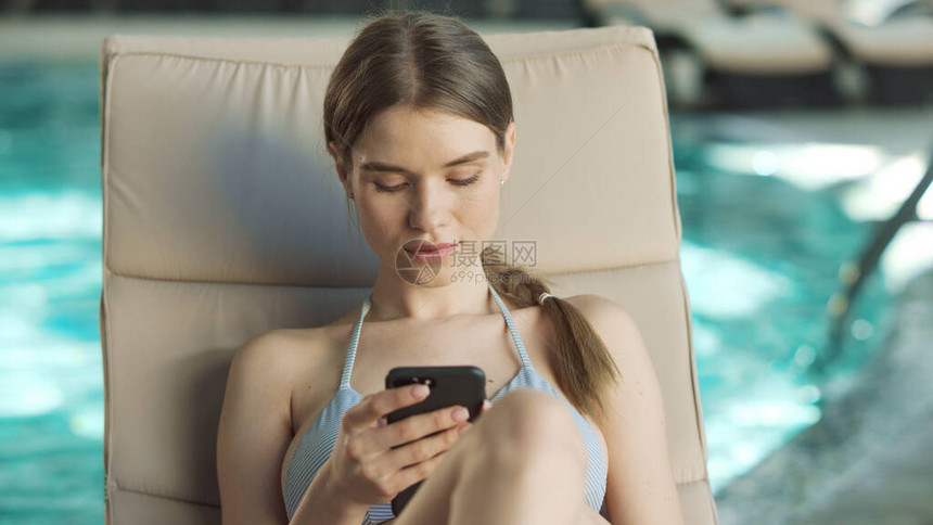 在豪华水疗中心游泳池边用电话放松的感女人的画像漂亮的女孩在躺椅上滚动手机与室内鸡尾酒年轻女子在酒店的电话图片