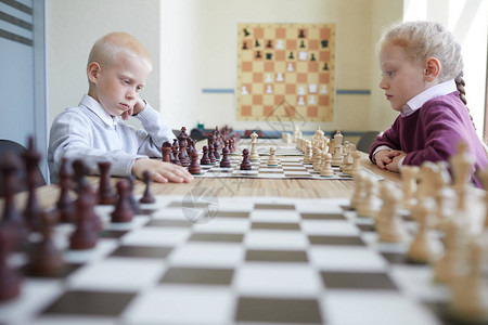 在国际象棋俱乐部教室的桌子上下棋的金发男孩和女孩图片