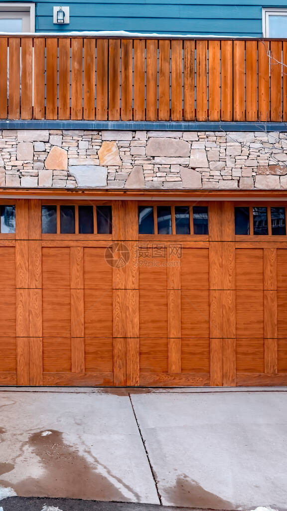 垂直作物棕色木制玻璃镶板车库门靠在家庭阳台下的石墙上在车道周围可以看到成堆的雪图片