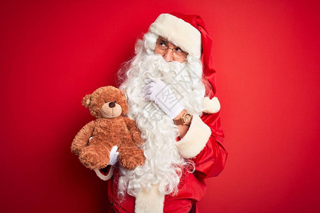 穿着圣诞老人服装的中年男子抱着泰迪熊在孤立的红色背景上严肃地思考问题图片