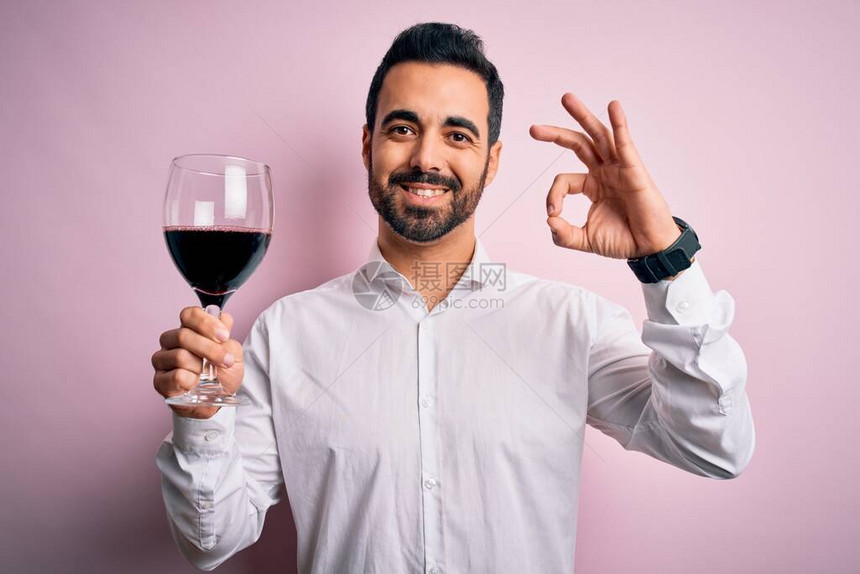长着胡子的英俊小帅哥喝着红酒杯子和孤立的粉红背景在用图片