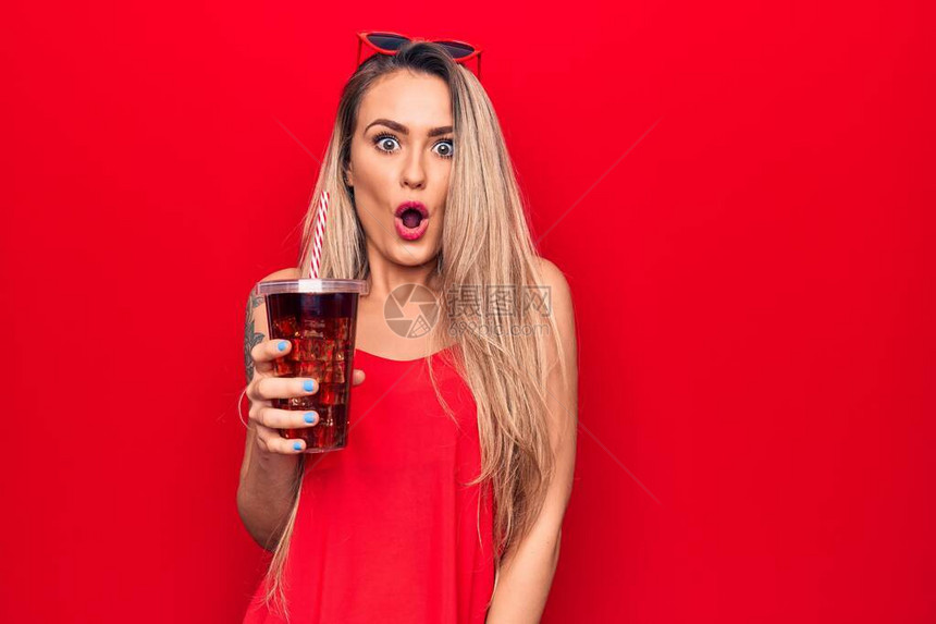 美丽的金发美女喝可口乐饮料的清酒用稻草在红色背景上吸干惊吓并图片