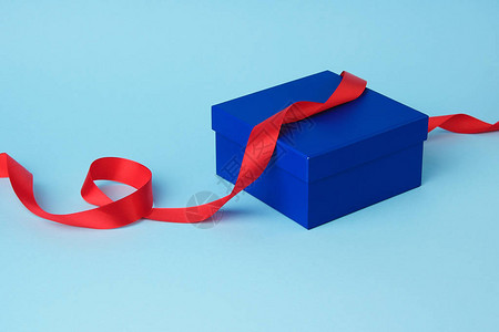 用于礼物的封闭方形蓝色纸板箱和蓝色背景上扭曲的丝绸红丝带背景图片