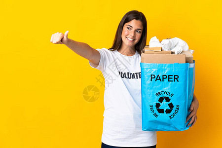拿着一袋废纸的回收袋来回收在黄色背景下被孤立的循环用拇指举起手势的图片