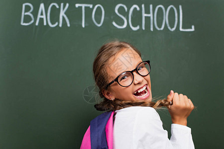 兴奋的女学生在眼镜里有选择焦点触摸头发在黑板边笑图片