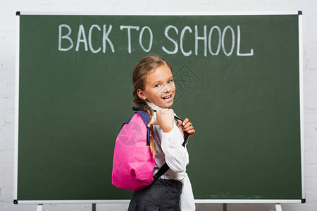 快乐的女学生用拇指着黑板附近的背包图片