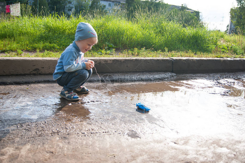 一个戴着蓝色帽子牛仔裤和凉鞋的三岁白人男孩在凉爽的夏日里开着一辆过时的遥控车玩耍孩子在绿草的背景下侧身图片