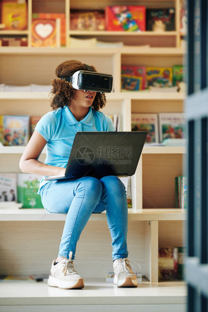 穿着休闲服的少女坐在现代学校图书馆使用VR眼镜和笔记本图片