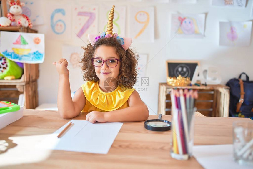 漂亮的蹒跚学步的孩子戴着眼镜和独角兽王冠坐在幼儿园的桌子上图片