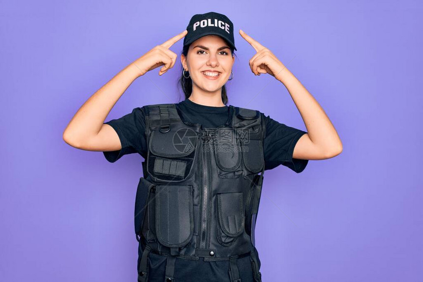 身穿紫色背景安全防弹背心制服的年轻女警用双手指着头微笑图片